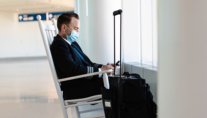 pilot sitting in rocking chair with face mask on next to luggage, looking own at cell phone device