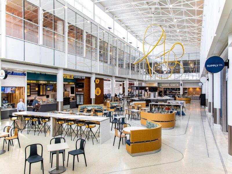 complete image of East Terminal Expansion, large café area with tables, chairs and restaurants, there is also art fixtures hanging from the ceiling  