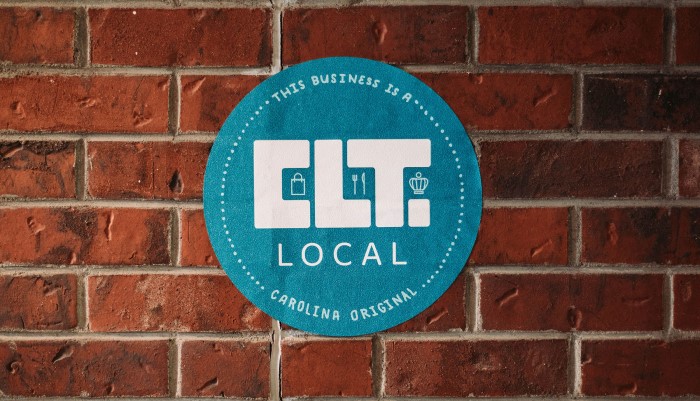 CLT Local signage posted on brick wall, sign is circle shape with teal background and white lettering