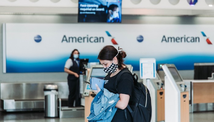 passenger featured in front of American Airline ticking counter, checking cellular device, wearing face mask and a backpacking carrying a jean jacket