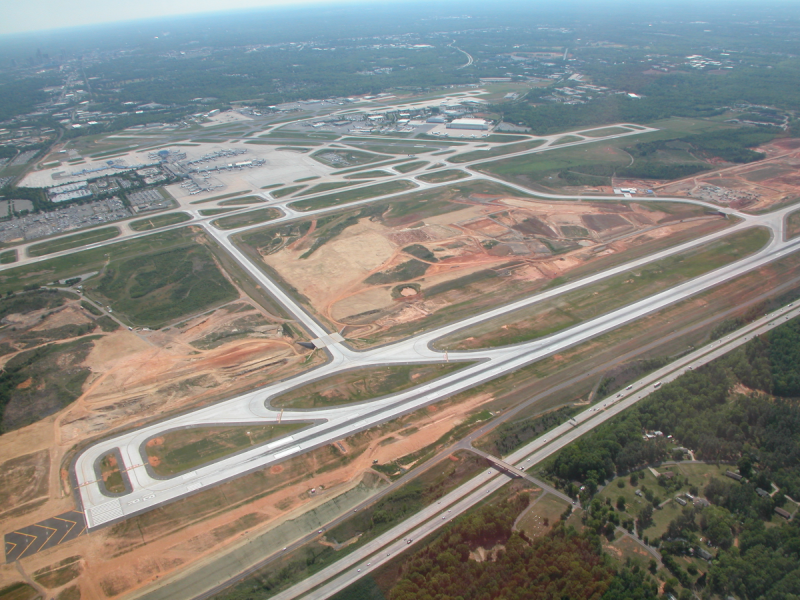 complete image of Third Parallel Runway - 18R/36L, dirt pathway with concrete outline of the runway, surrounded by trees, airport terminal and buildings