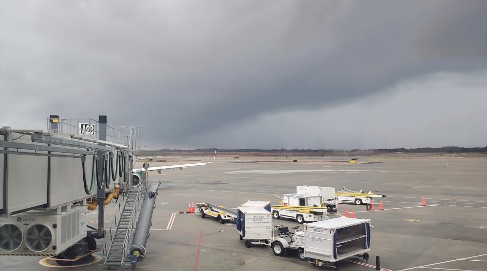 Storny weather overlooking the airfield. A grounded plane at the jetbridge and tugs parked nearby. A large tornado is visible on the horizon.