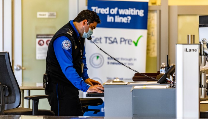 TSA employee on the phone and typing on the computer at workstation; employee is wearing mask and TSA uniform, in background is TSA Pre-check promotional banner