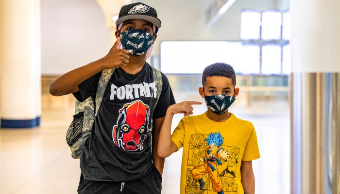 two young boy passengers wearing face masks and posing with thumbs up, featured in terminal lobby, wearing graphic t-shirts