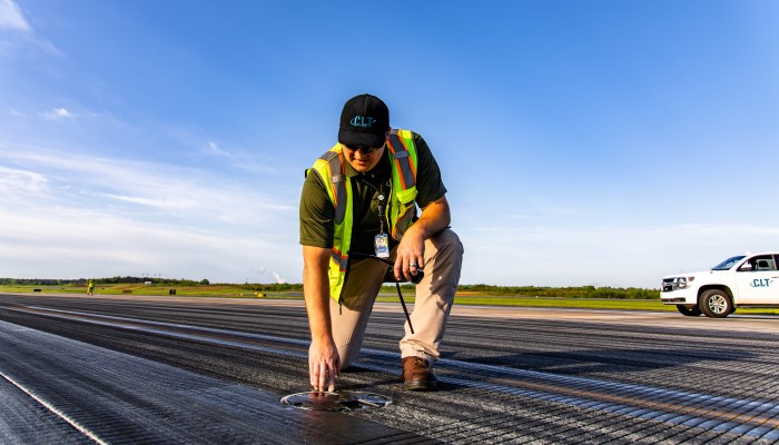 CLT Airport employee featured on airfield checking ground levels; employee is wearing CLT branded apparel and safety vest; clear blue sky with CLT work truck in background