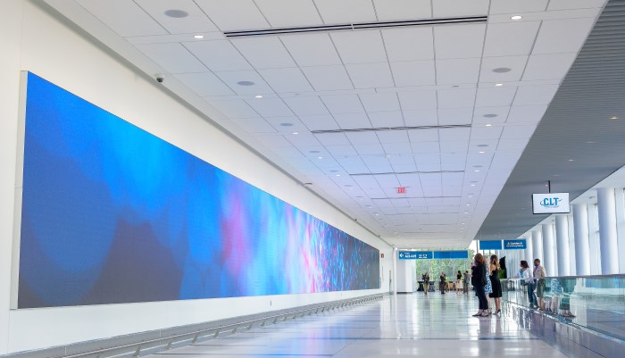Passengers featured standing in front of a digital mural in Concourse A near moving sidewalk; mural has colors of blue, pink and purple