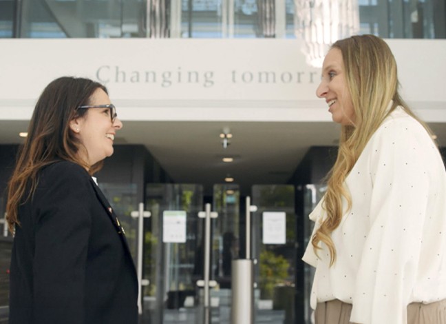 「明日は変えられる。」と書かれた建物の前で、二人の女性が笑顔で向かい合っています。