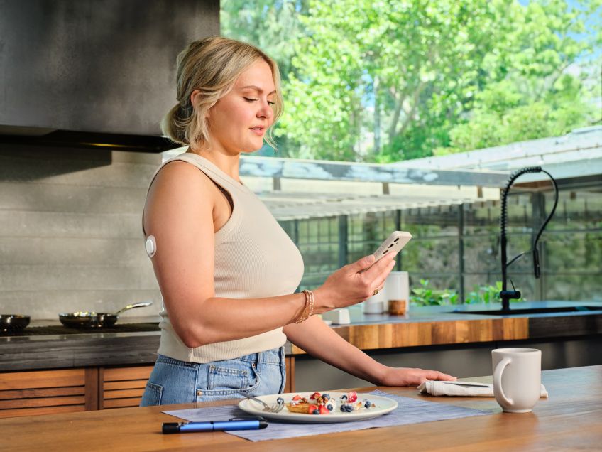 A woman in a kitchen wearing a Simplera sensor on her arm and checking her phone. 